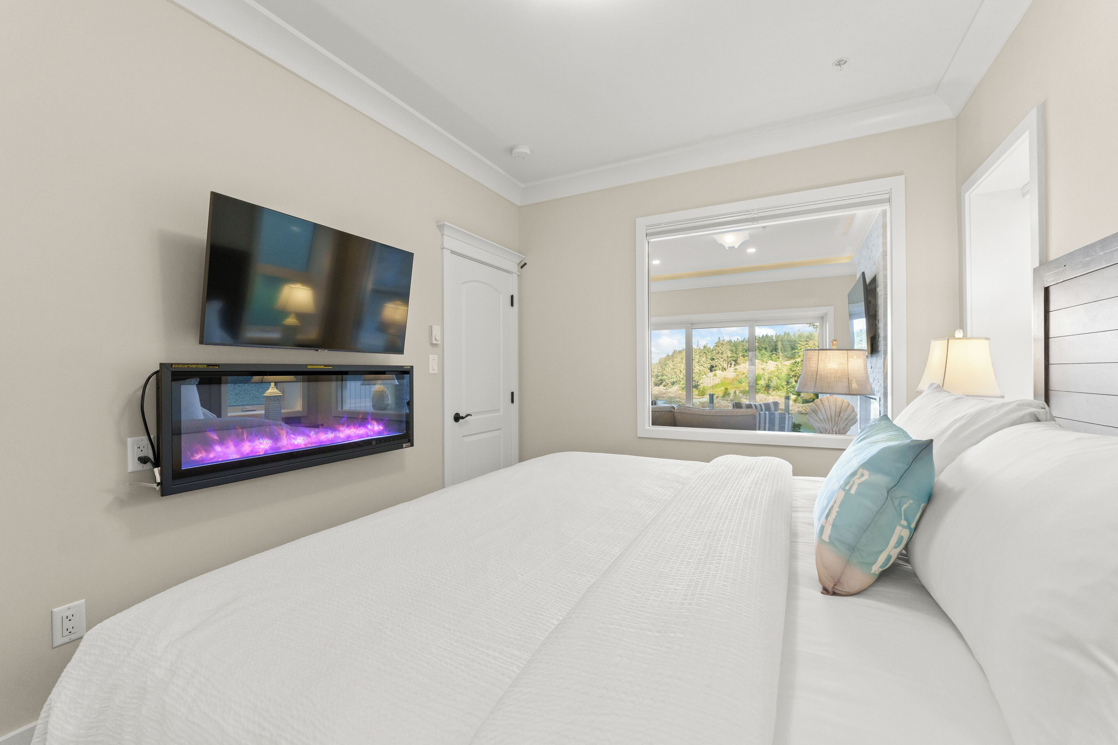 A hotel room with a large bed, a mounted TV, a window with a view of greenery, and a wall-mounted aquarium.