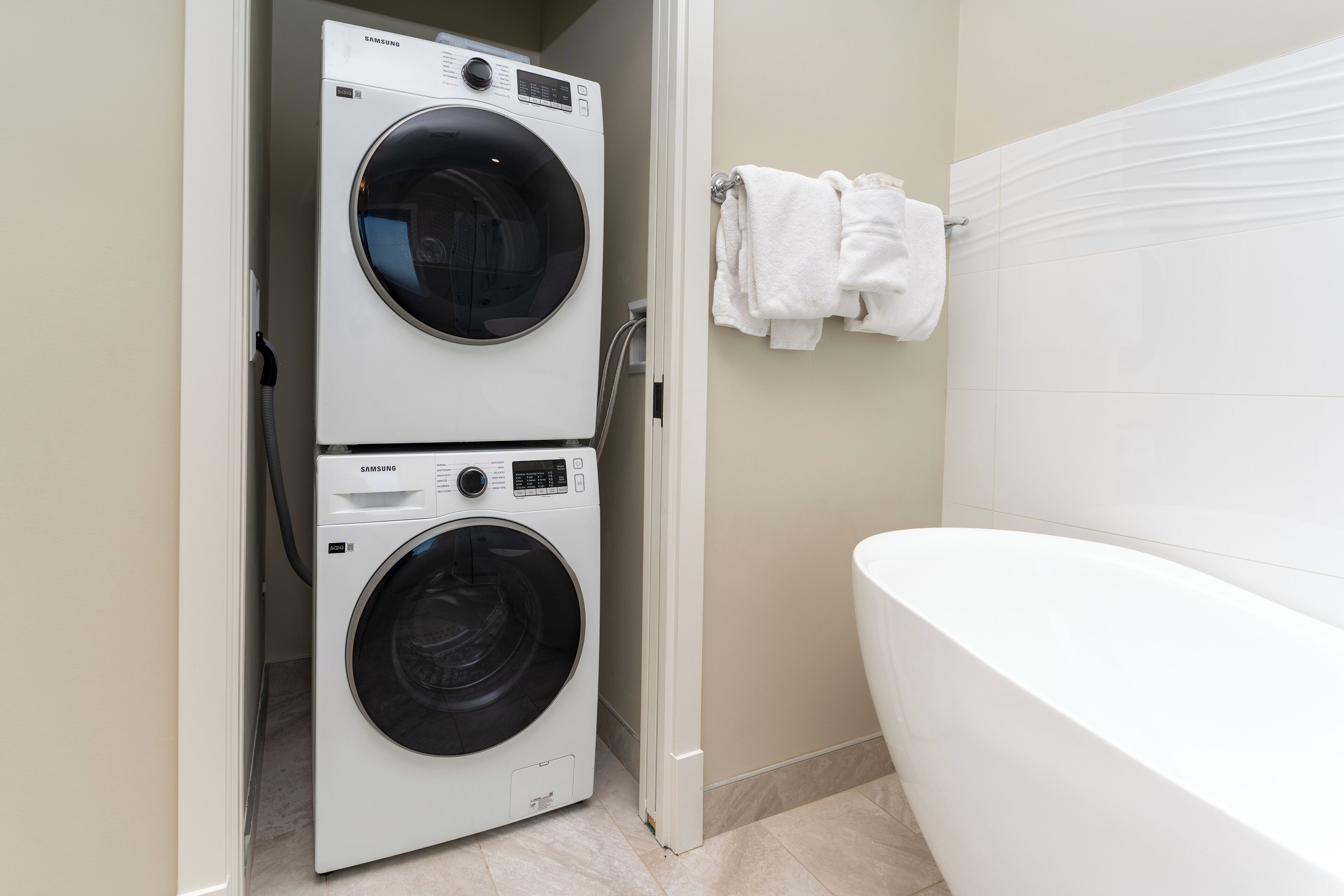 Un cuarto de lavado con lavadora y secadora de carga frontal, una bañera blanca y un perchero con toallas dobladas.