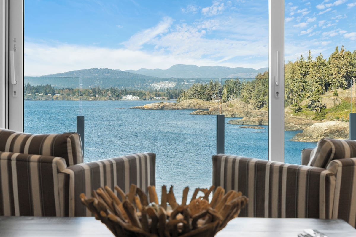 A room with a large window overlooking a lake, two striped armchairs, and a wooden decorative piece on the table.