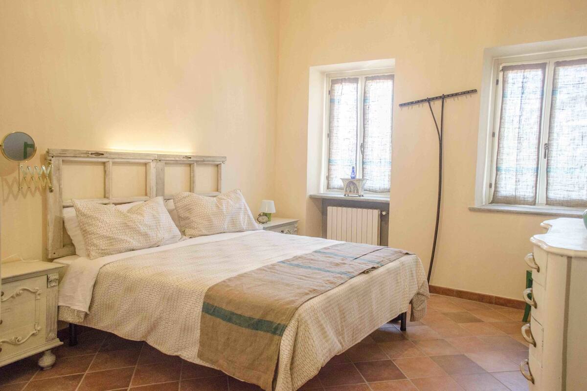 Un dormitorio con una cama, mesitas de noche, cómoda, radiador y dos ventanas con cortinas.