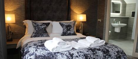 A hotel room with a large bed, two bedside lamps, a bathroom with a sink and mirror, and a glass door leading to a shower area.
