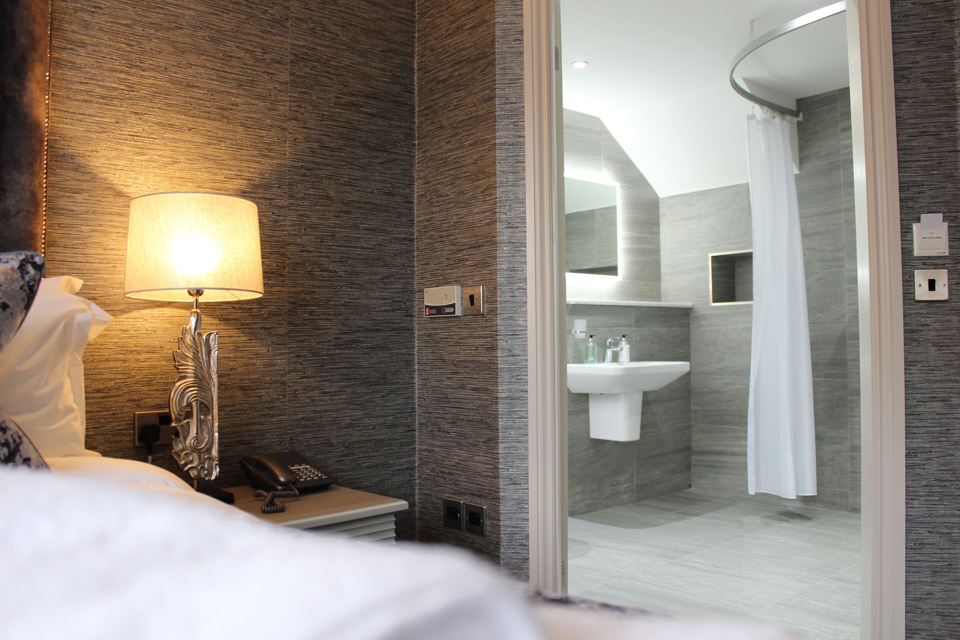 A hotel room with a bed, a bedside lamp, a telephone, and a view into the bathroom through a glass door.