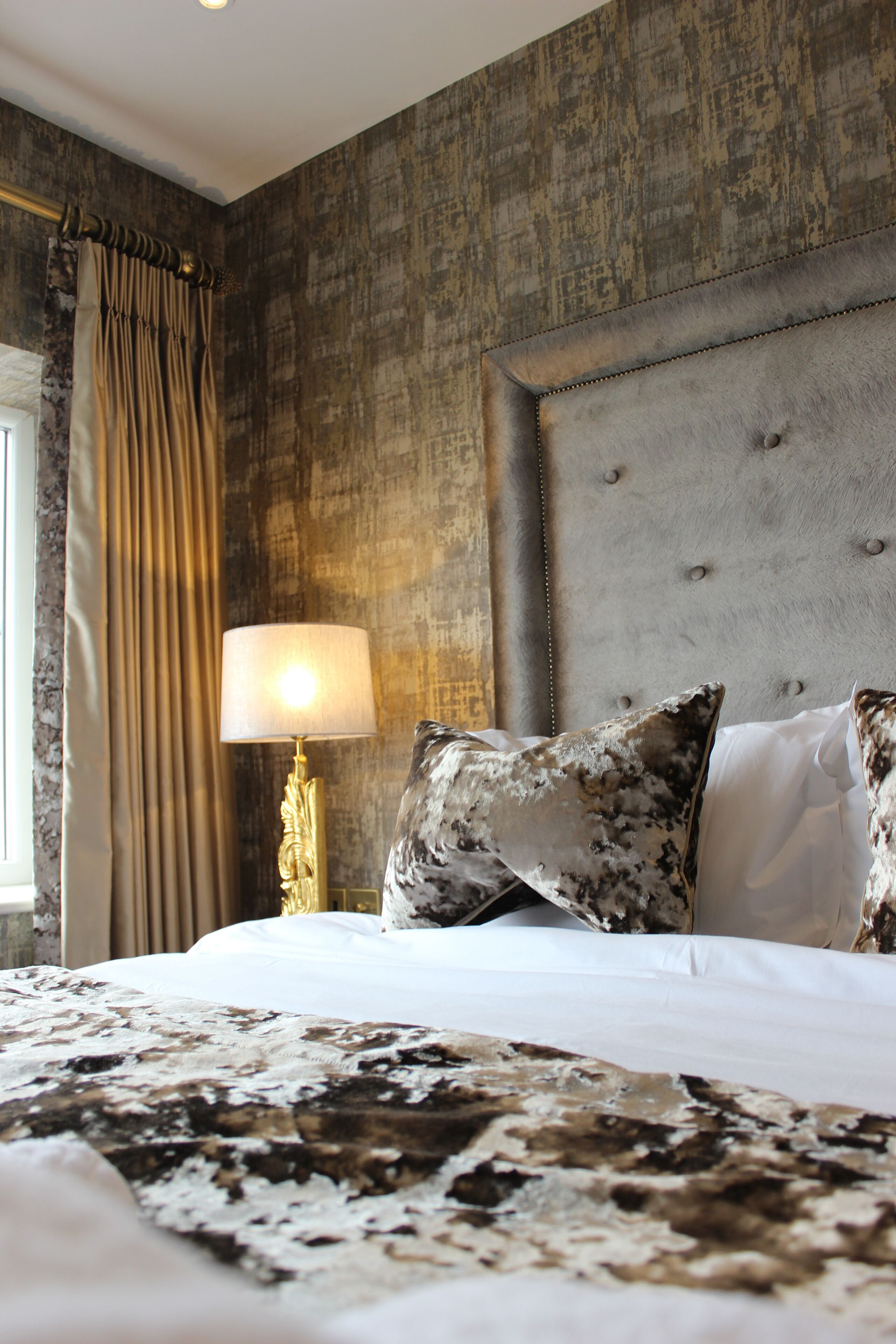 A bedroom with a tufted headboard, a bed with patterned bedding, a bedside lamp, and curtains.
