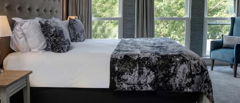 Un dormitorio con una cama grande, cabecera, mesita de noche, una silla y una ventana con cortinas.