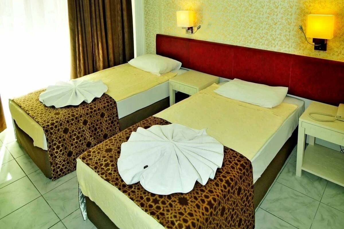 A hotel room with two beds, each with a red headboard, white linens, and a giraffe-patterned bedspread. There are two bedside lamps, a telephone, and an air conditioning unit on the wall.