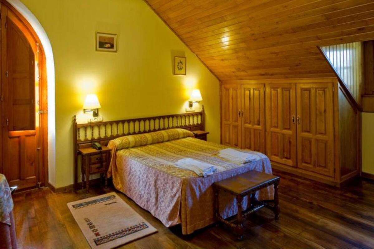 A bedroom with a wooden ceiling, a bed with a patterned bedspread, two bedside lamps, a wooden wardrobe, and a small bench.