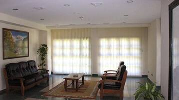 Amplia sala de estar con un sofá de cuero oscuro, una mesa de centro de madera y dos sillones. Se aprecia un ventanal con persianas verticales y una planta en maceta a la derecha.