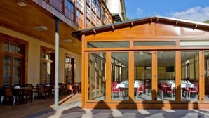 Un café al aire libre con mesas y sillas de madera, grandes ventanales de vidrio y piso de baldosas.