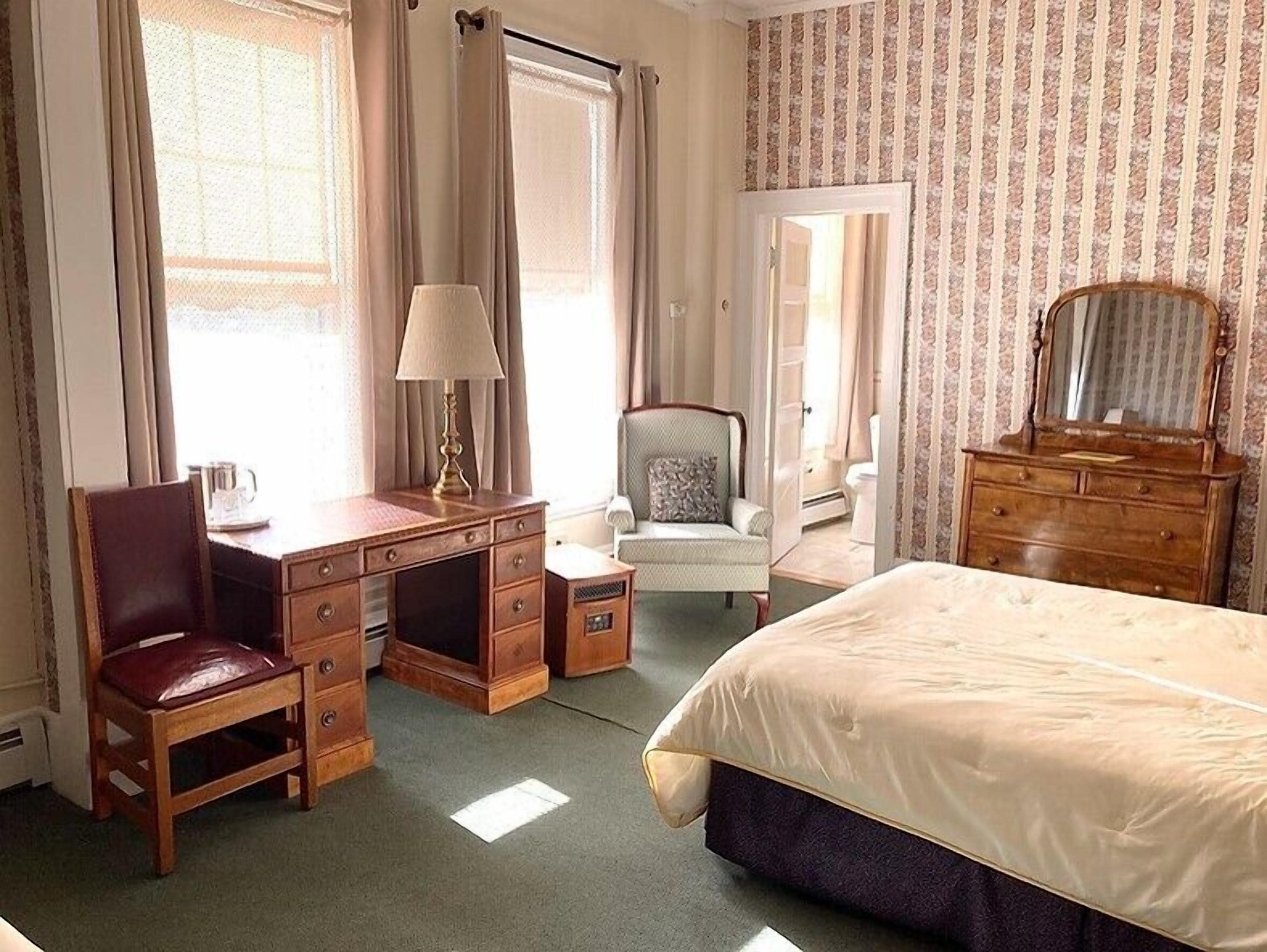 A hotel room with a bed, a desk with a chair, a dresser, and a chair with a pillow.