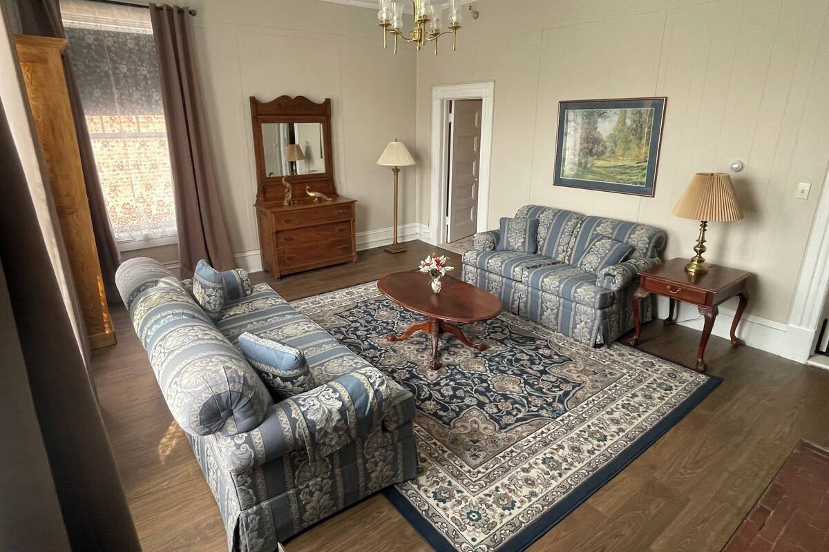A living room with a patterned sofa, a matching armchair, a coffee table, a wooden dresser, a mirror, a lamp, and a painting on the wall.