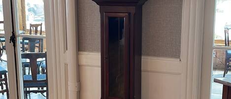 A tall, dark wooden grandfather clock with a square face and Roman numerals, placed in a room with wooden flooring and white trim.