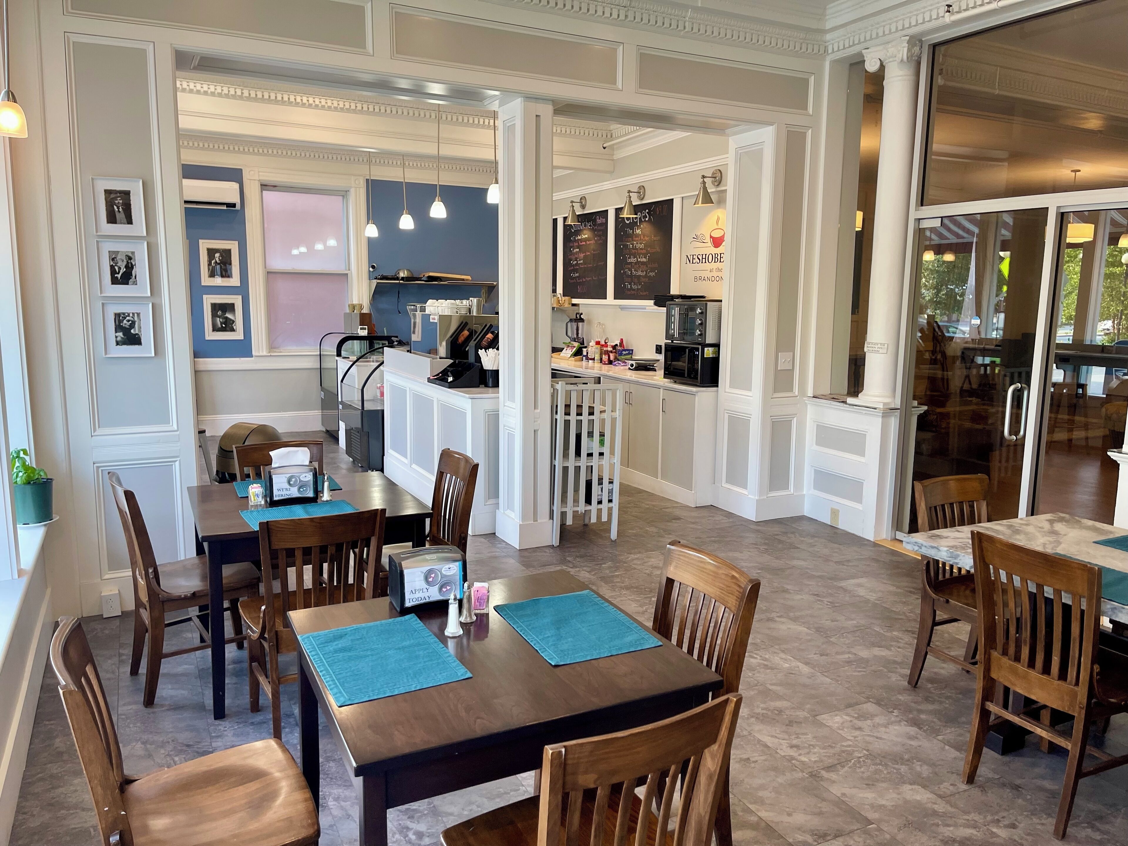 A dining area with wooden tables and chairs, a kitchen in the background, and framed pictures on the wall.