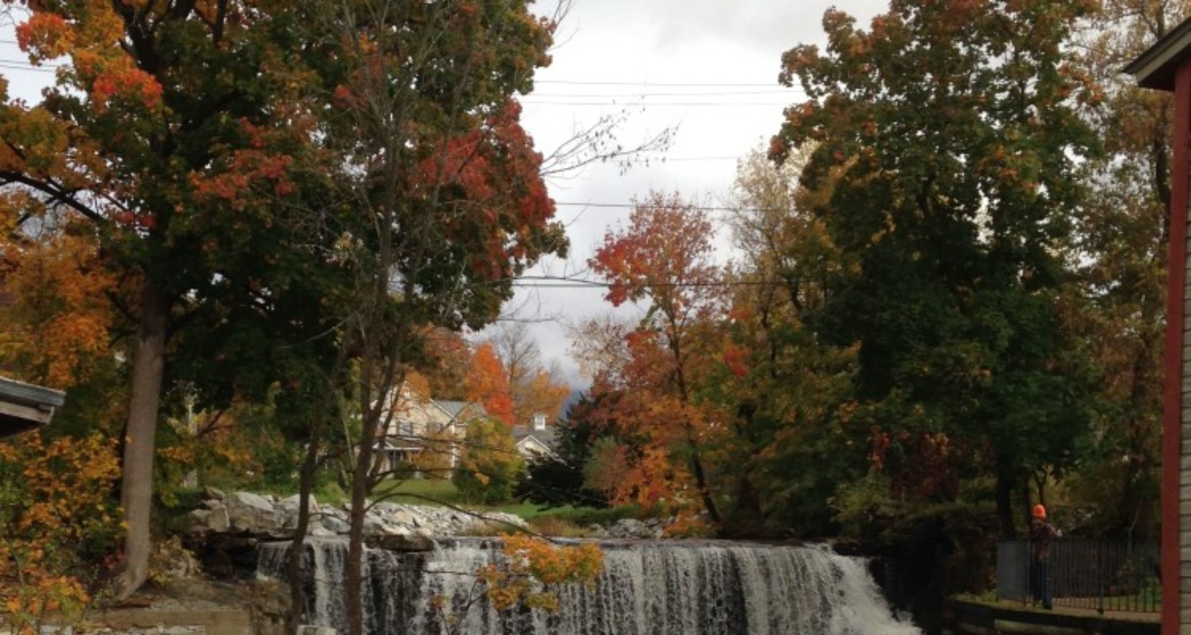 A waterfall surrounded by trees with autumn foliage, a stone bridge, and a house in the background.