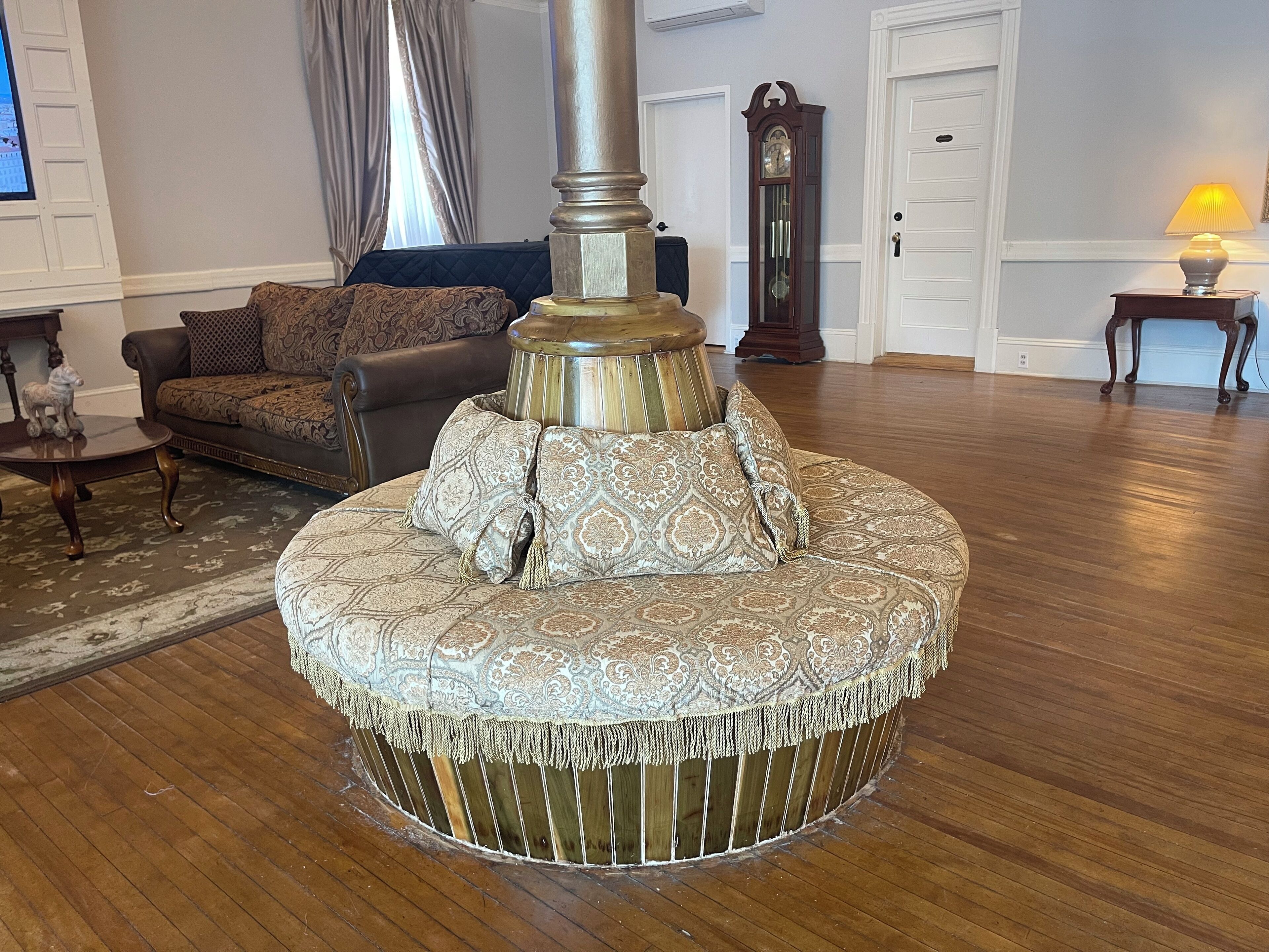 A round, upholstered ottoman with a decorative base, positioned in a room with wooden flooring, a sofa, a table, and a lamp.