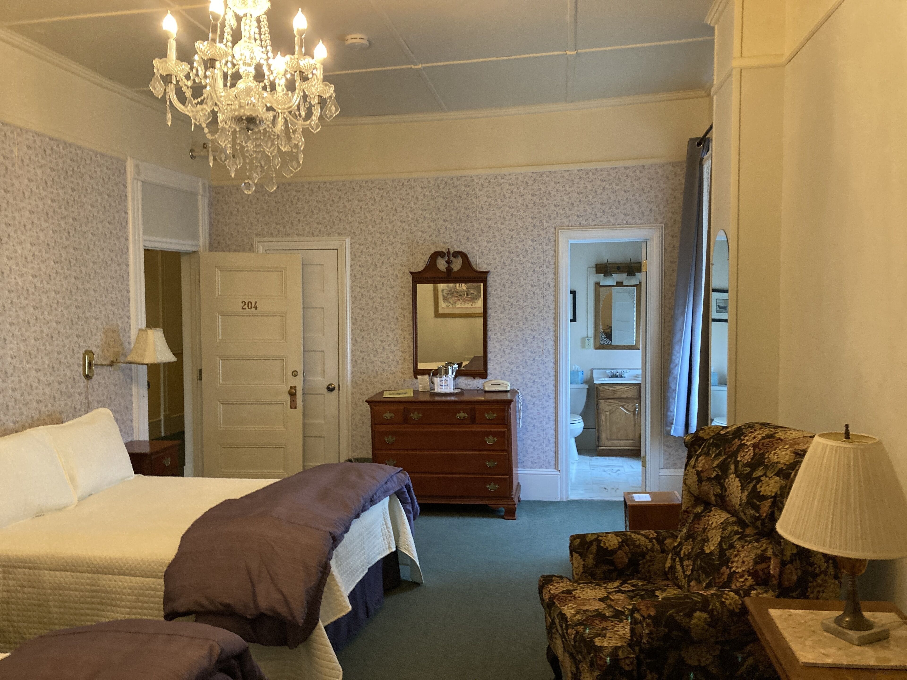 A hotel room with a bed, a dresser, a chair, and a chandelier.