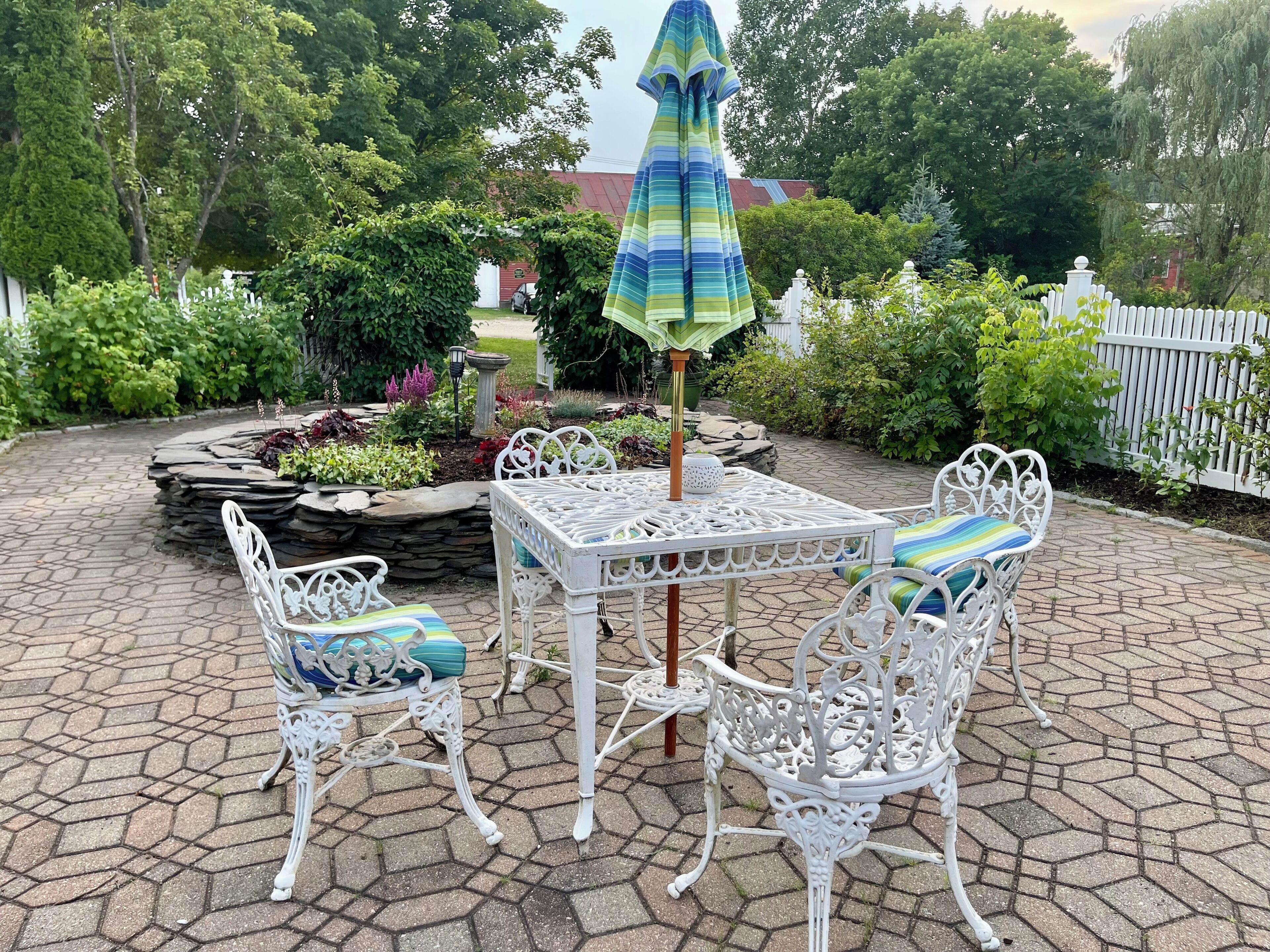 Une terrasse avec une table et des chaises en métal blanc, entourée d’un jardin délimité par une clôture en bois peinte en blanc.