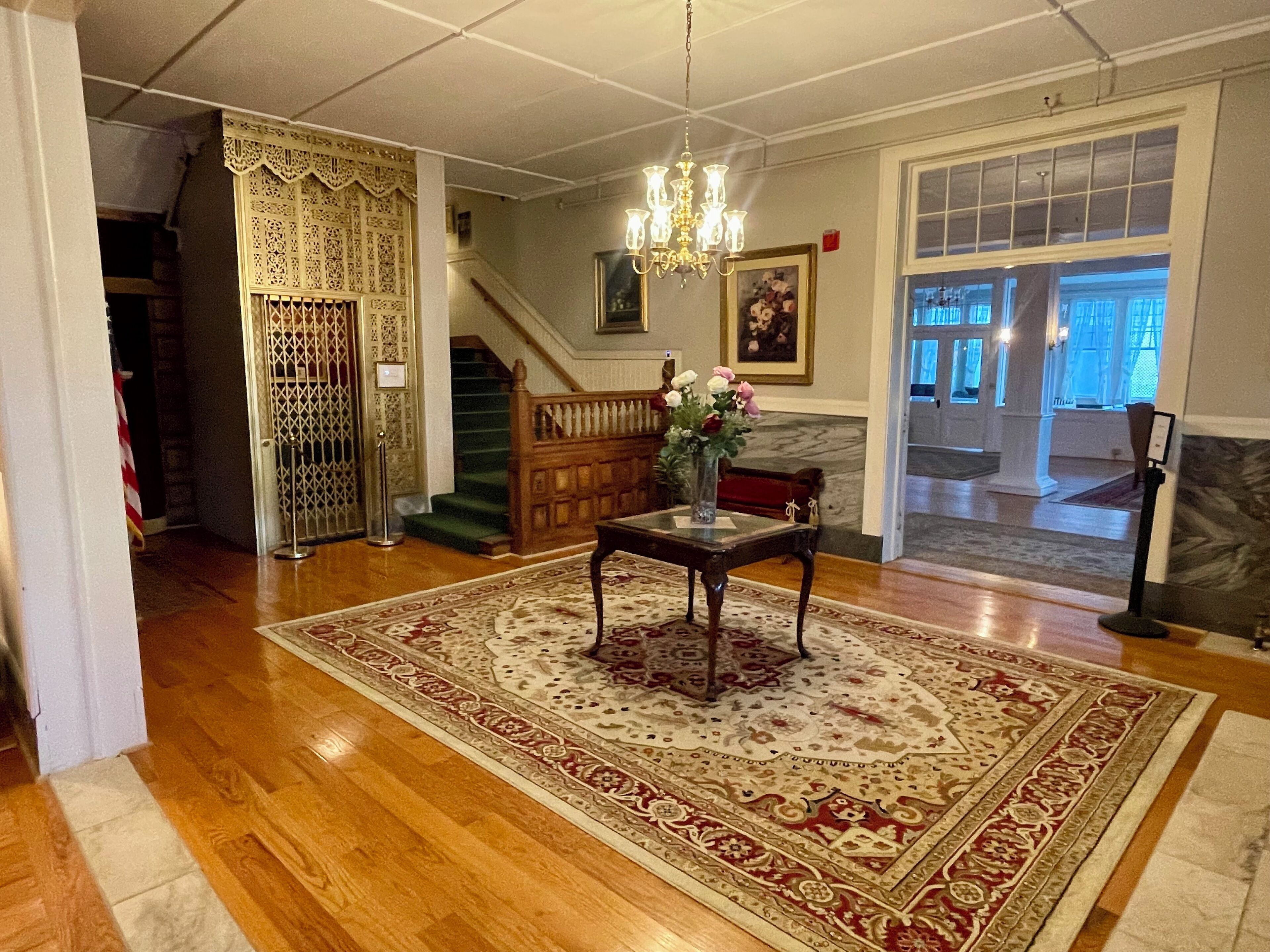 A spacious living room with hardwood floors, a large ornate rug, a wooden coffee table with a vase of flowers, a chandelier, and a staircase leading to another level.