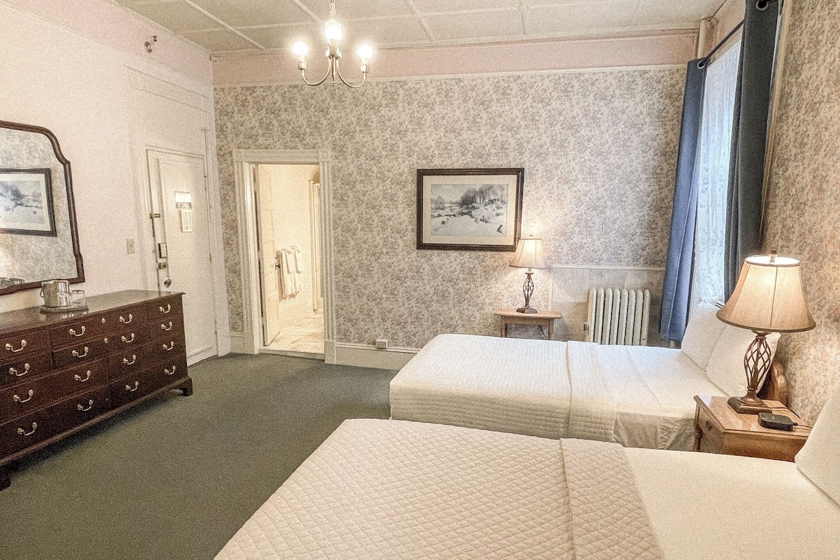 A hotel room with two beds, a dresser, a chandelier, a framed picture, and a radiator.