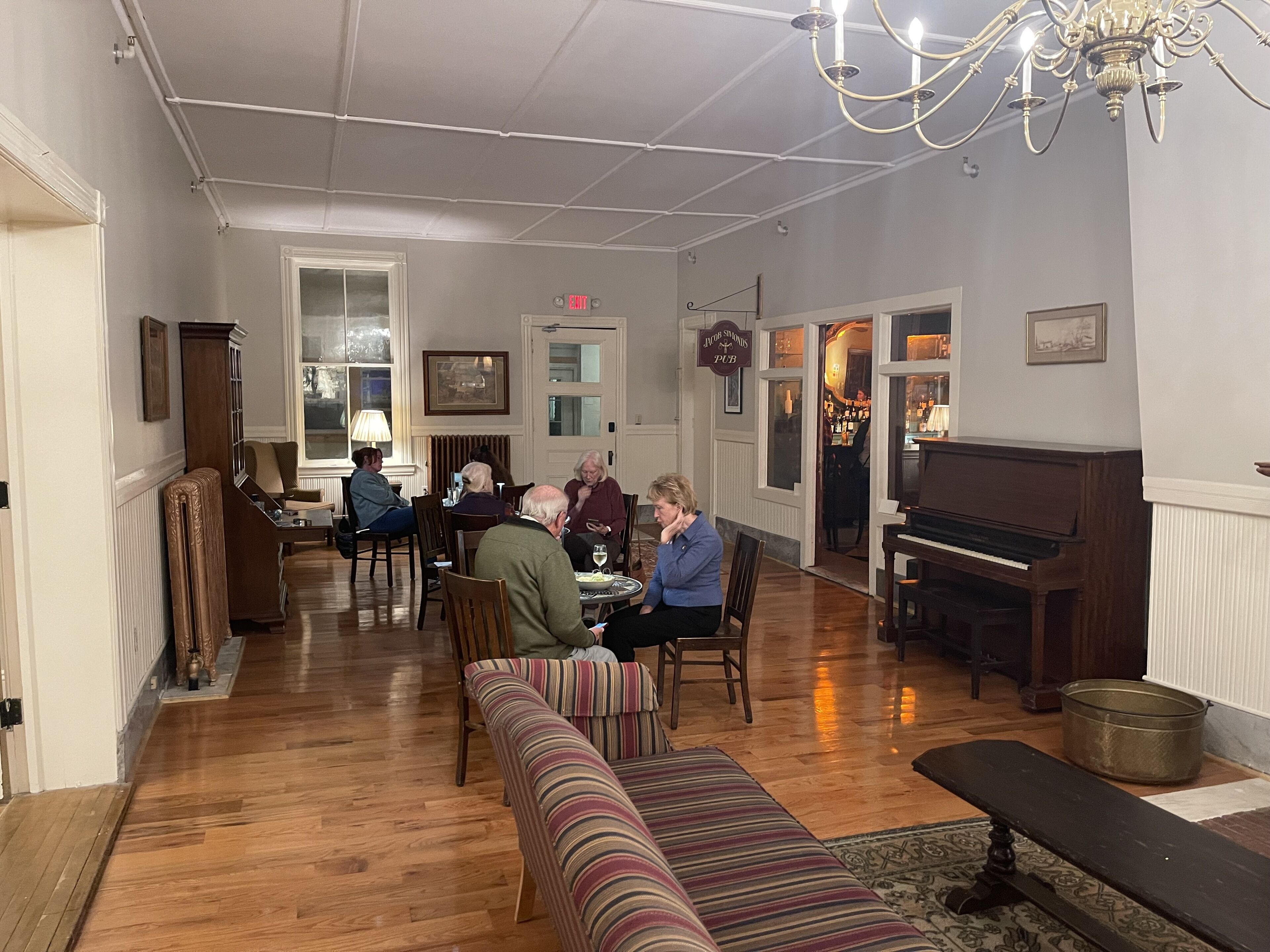 Une pièce chaleureuse avec un plancher en bois, un piano, un banc et un groupe de personnes assises autour d’une table.