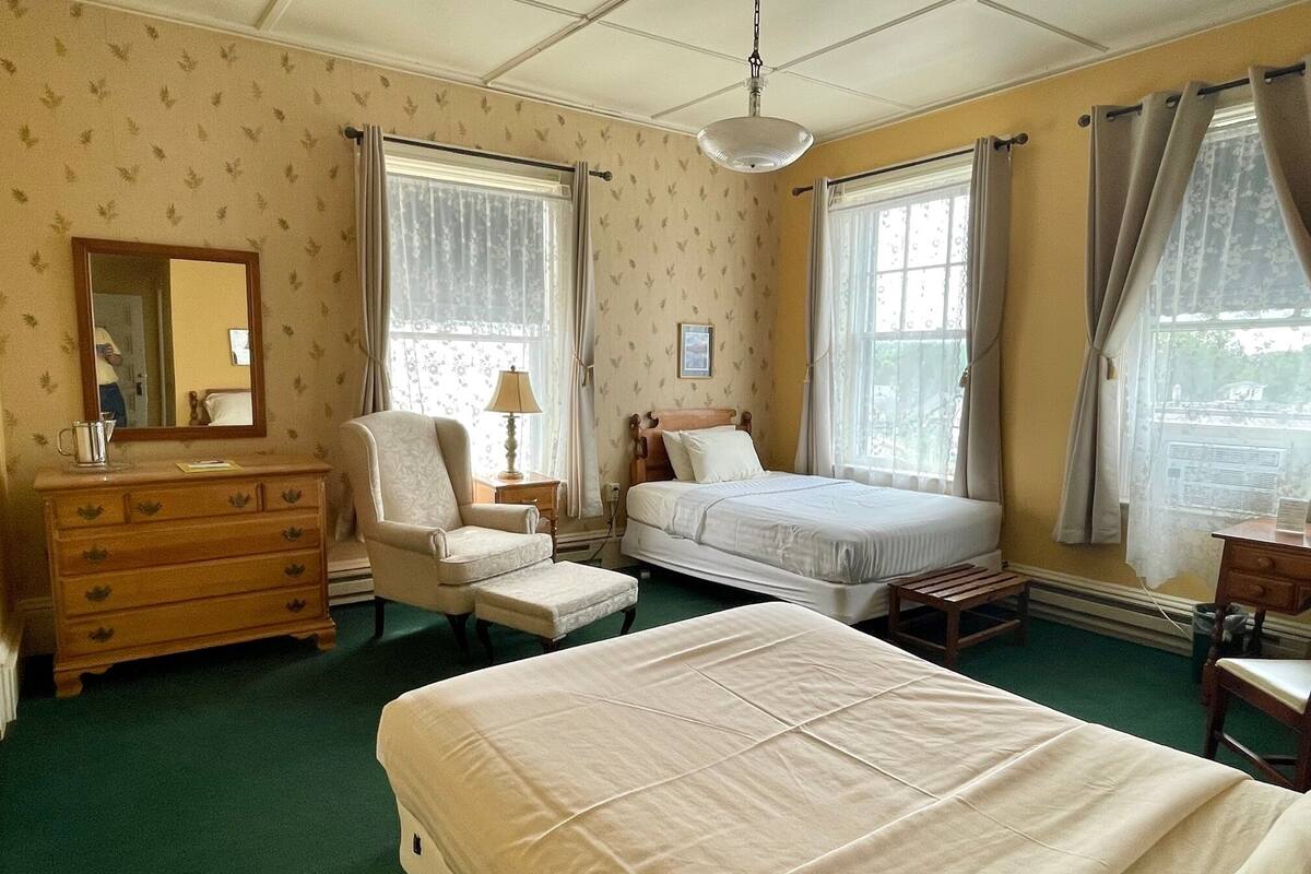 A hotel room with a bed, a chair, a dresser, a mirror, and windows with curtains.