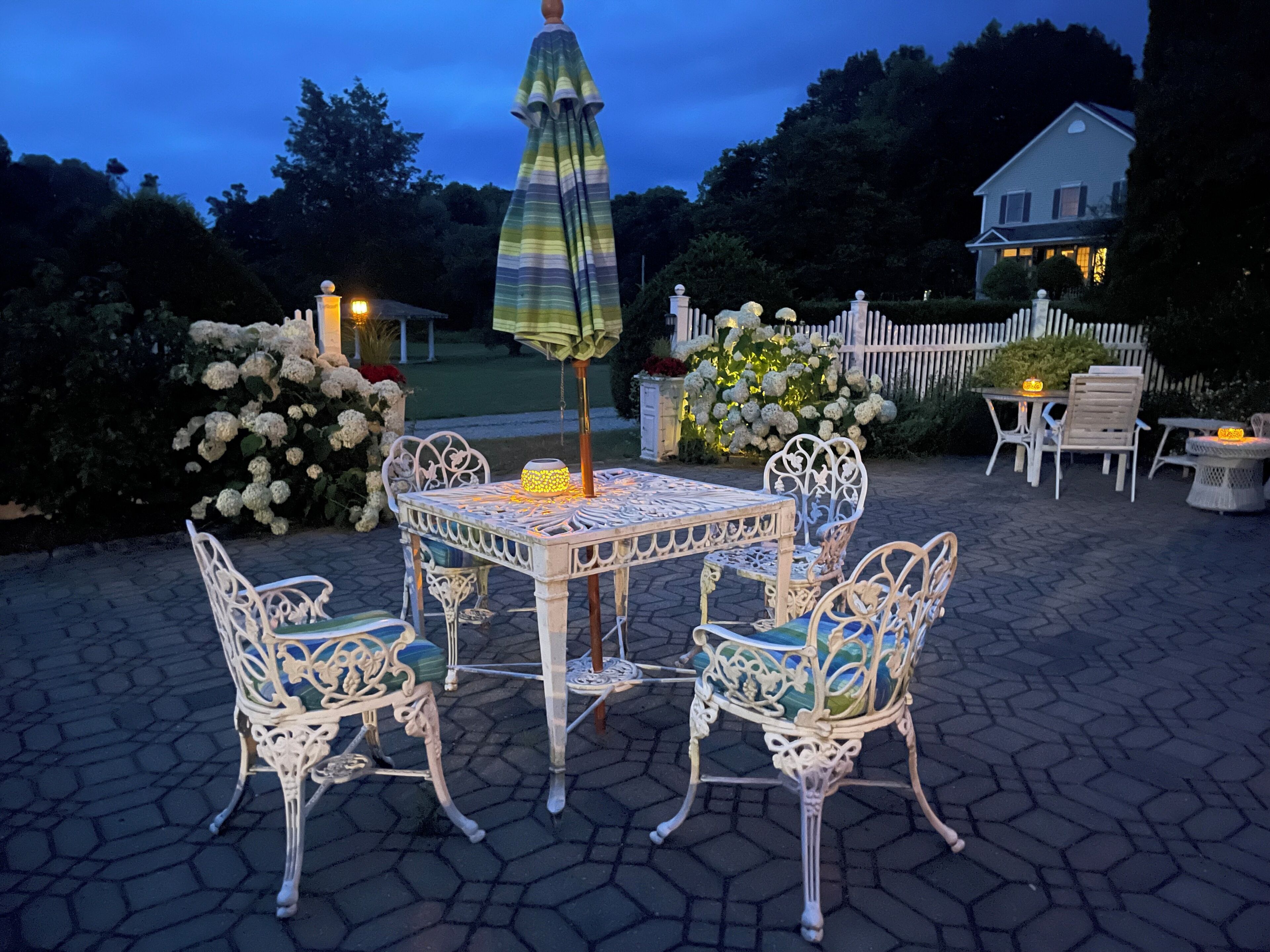 En udendørs terrasse med et bord og stole, omgivet af et hvidt hegn og et hus i baggrunden.