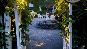 Una entrada al jardín con un pérgola blanca cubierta de hiedra, un letrero que indica que la piscina está abierta y mobiliario exterior con sombrillas.