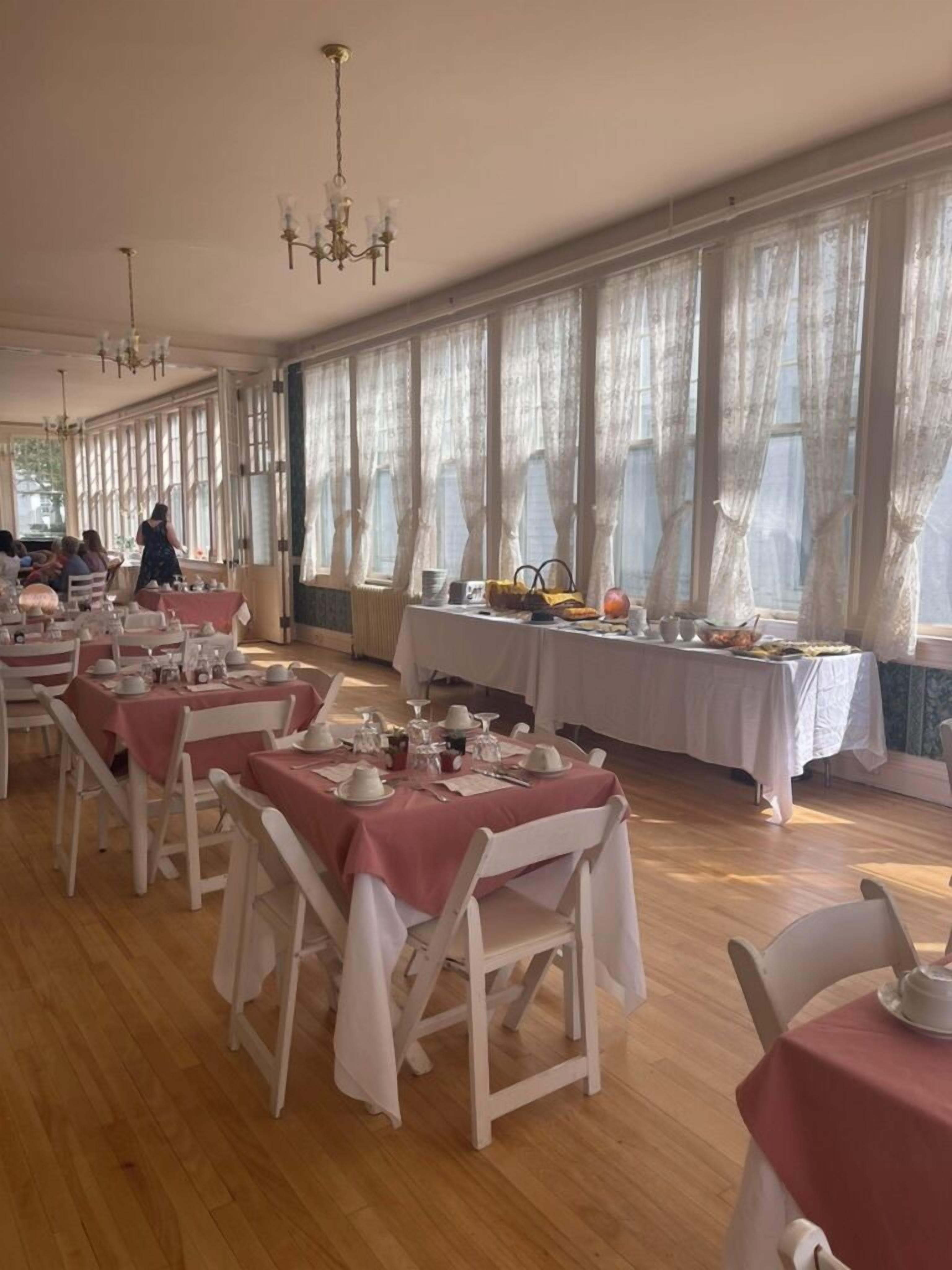 A dining area with tables set for a meal, large windows with curtains, and chandeliers.