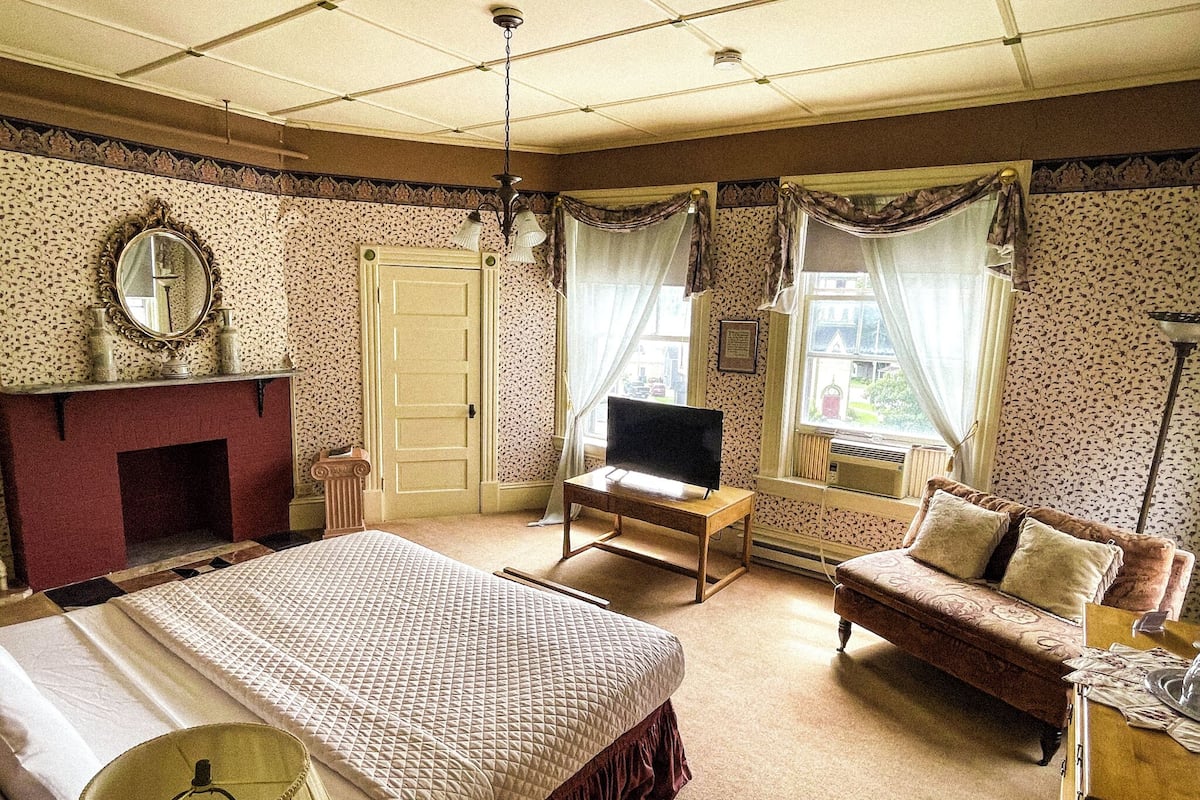 A bedroom with a bed, a television, a sofa, a fireplace, and two windows with curtains.