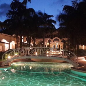 A nighttime view of a pool area with a bridge, palm trees, and a building with arched windows. - Tama's Villas (Malindi)