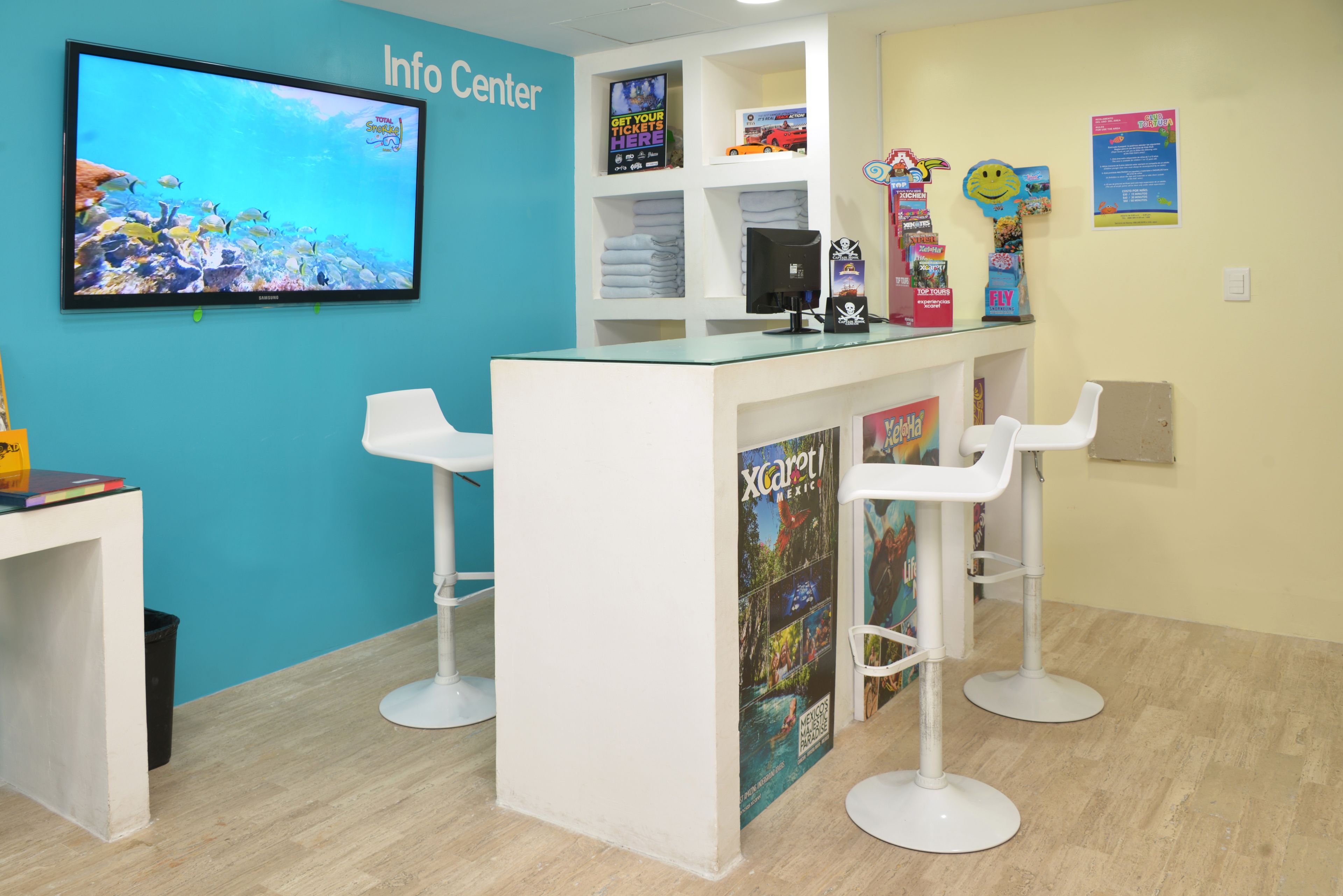 Ein Empfangsbereich mit einem Fernseher, der eine Meereslandschaft zeigt, einem Schild mit der Aufschrift 'Info Center' und einem Poster für 'Xcaret Mexiko'.