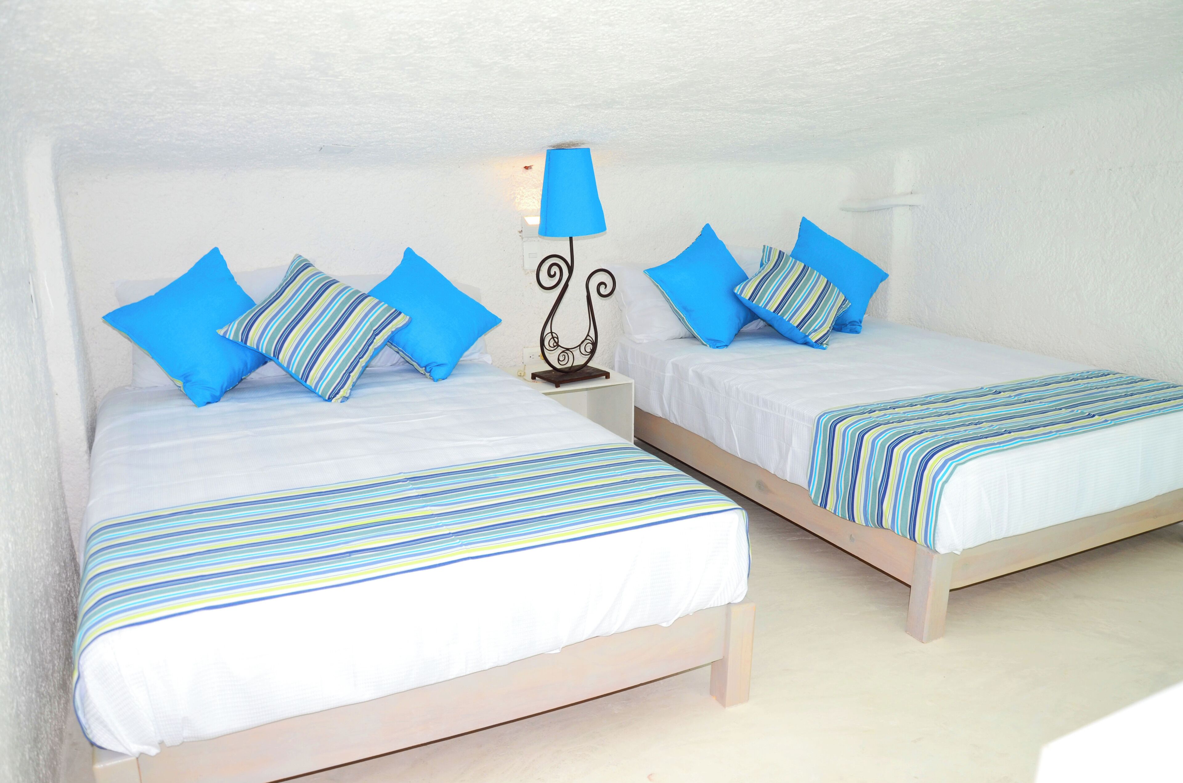 Ein Zimmer mit zwei Betten, jeweils mit einer gestreiften Tagesdecke und blauen Kissen. Dazwischen befindet sich ein Nachttisch mit einer Lampe.
