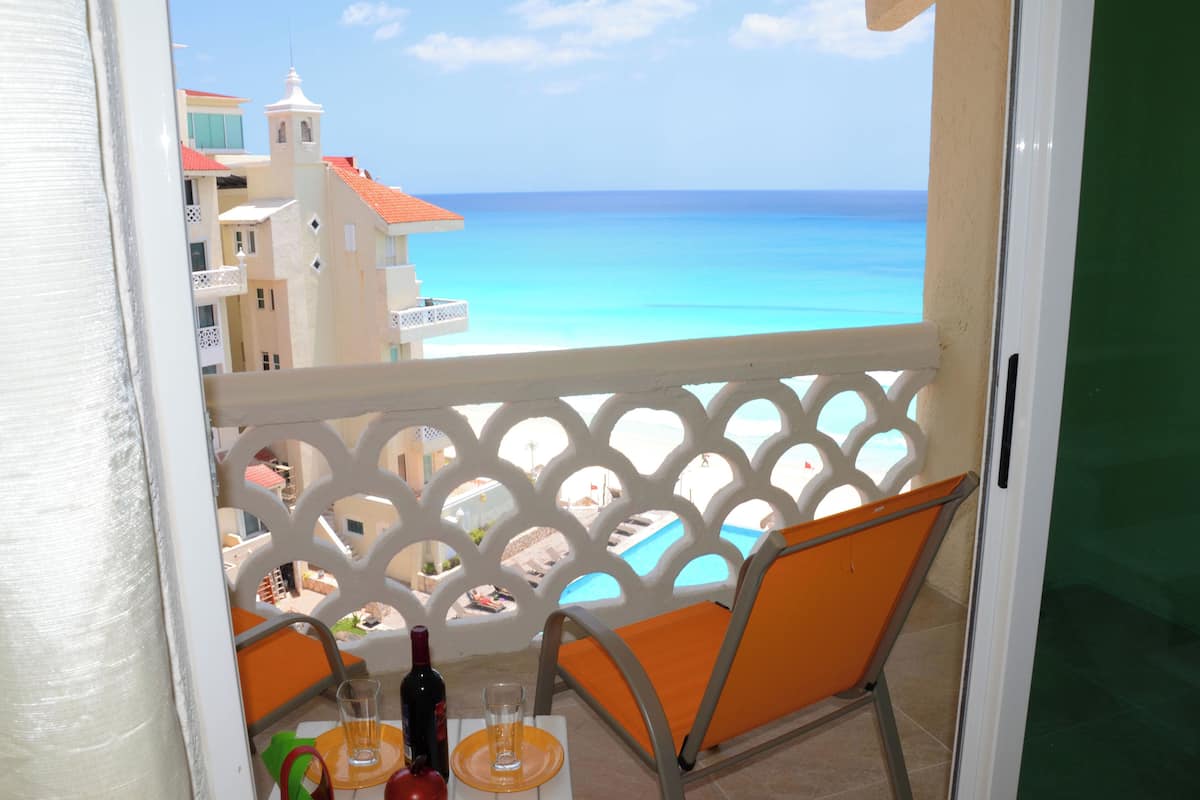 A balcony with a view of the ocean and buildings. There is a table with a bottle and glasses.