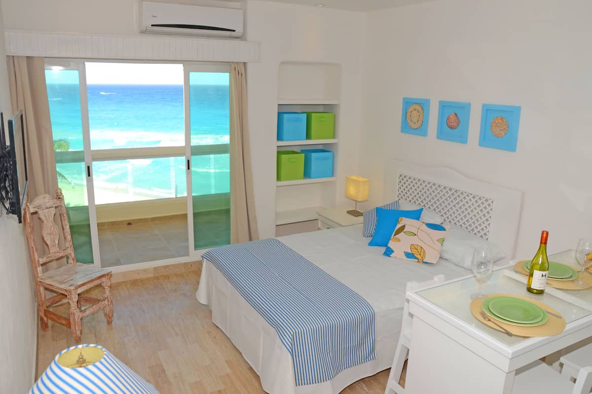 A hotel room with a bed, a chair, a table with a bottle and glasses, and a view of the ocean through a sliding door.