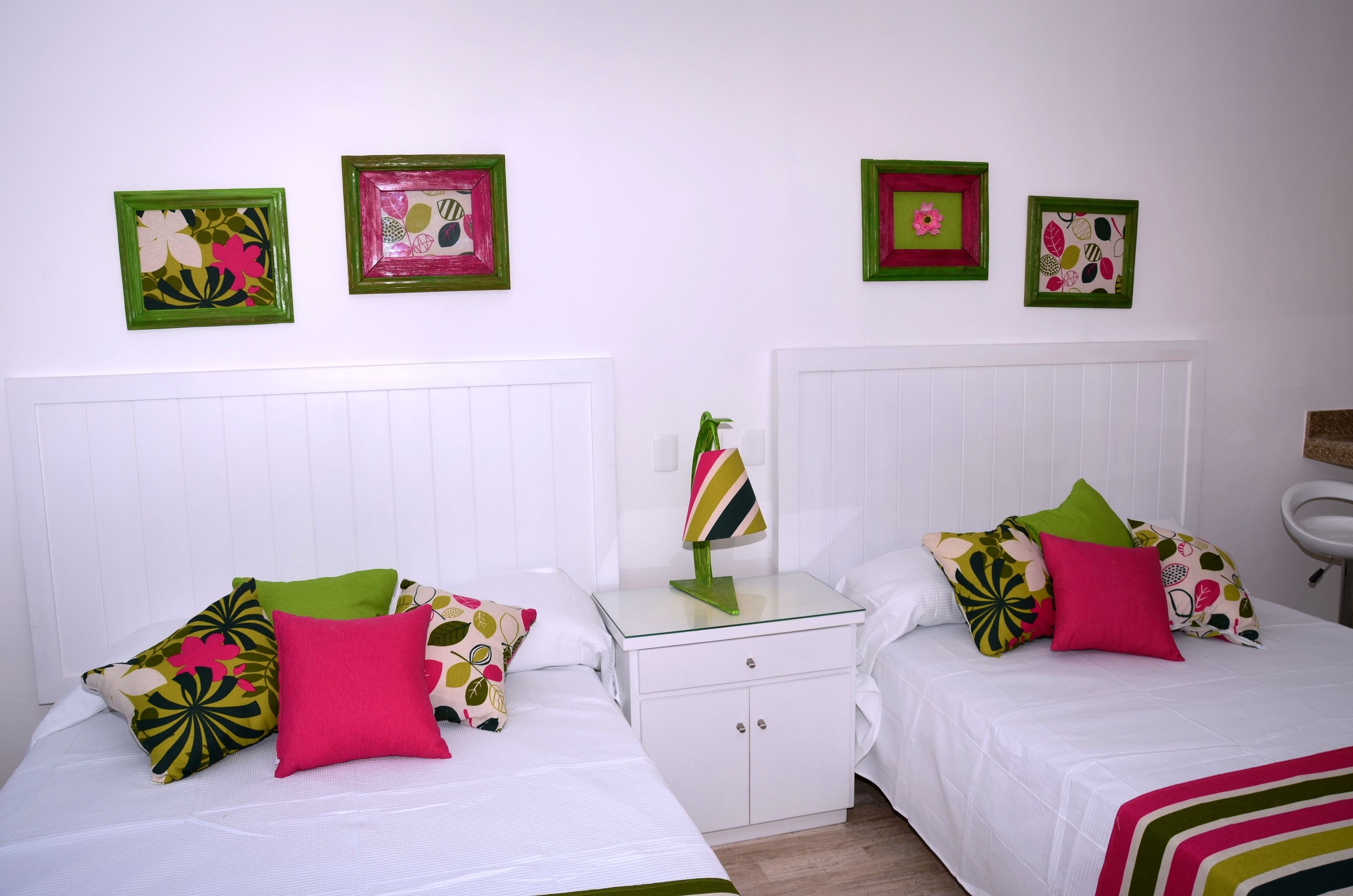Habitación de hotel con dos camas, cada una con almohadas estampadas, una mesita de noche con lámpara y cuadros en las paredes.
