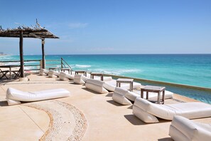 Un área de descanso en la playa con asientos blancos acolchados y una mesa de madera, con vista al océano.