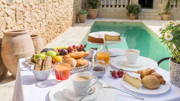 A table set with breakfast items including croissants, fruit, and a cake, with a pool in the background. - Hotel Casa Peya - Adults Only (Palafrugell)