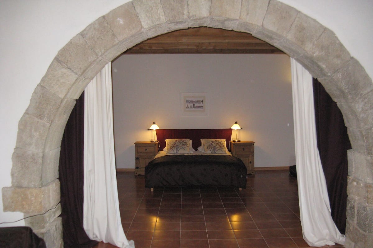 Une chambre avec une arche en pierre, un lit doté d  ’une tête de lit sombre, deux tables de chevet avec des lampes et un panneau accroché au mur.
