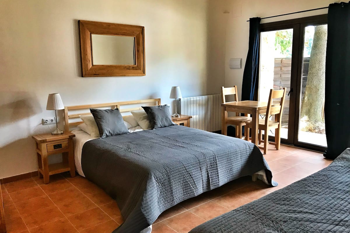 Une chambre à coucher comprenant un lit, des tables de chevet, une chaise, une table et une porte-fenêtre coulissante donnant sur un espace extérieur.