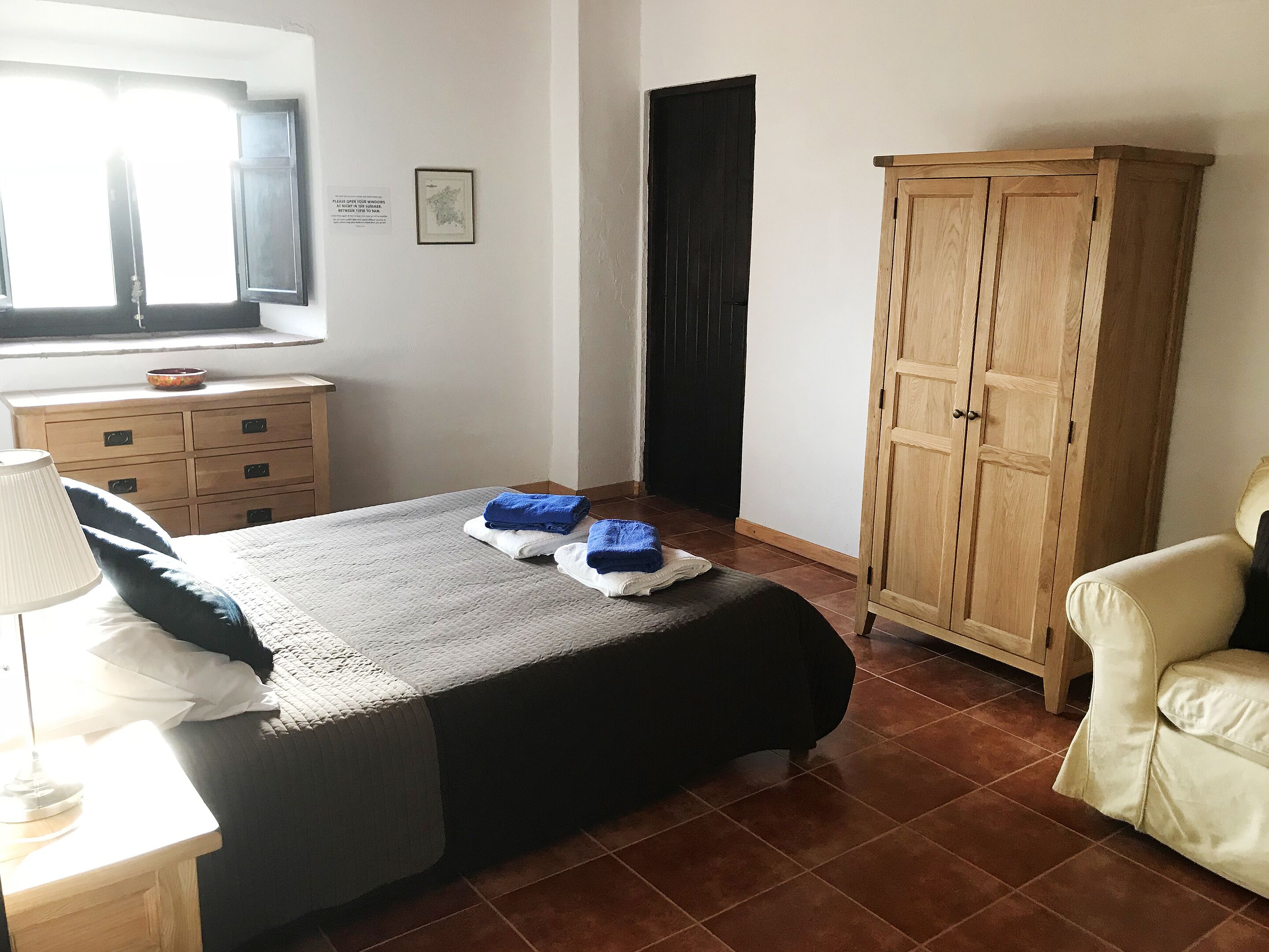 A bedroom with a bed, a wooden wardrobe, a desk with a lamp, and a window with blinds.