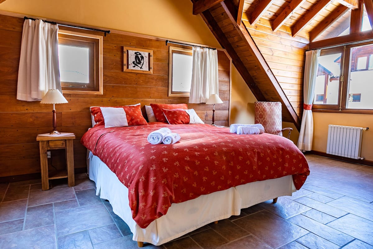 A bedroom with a wooden ceiling, a bed with red bedding, two bedside lamps, and a window with curtains.