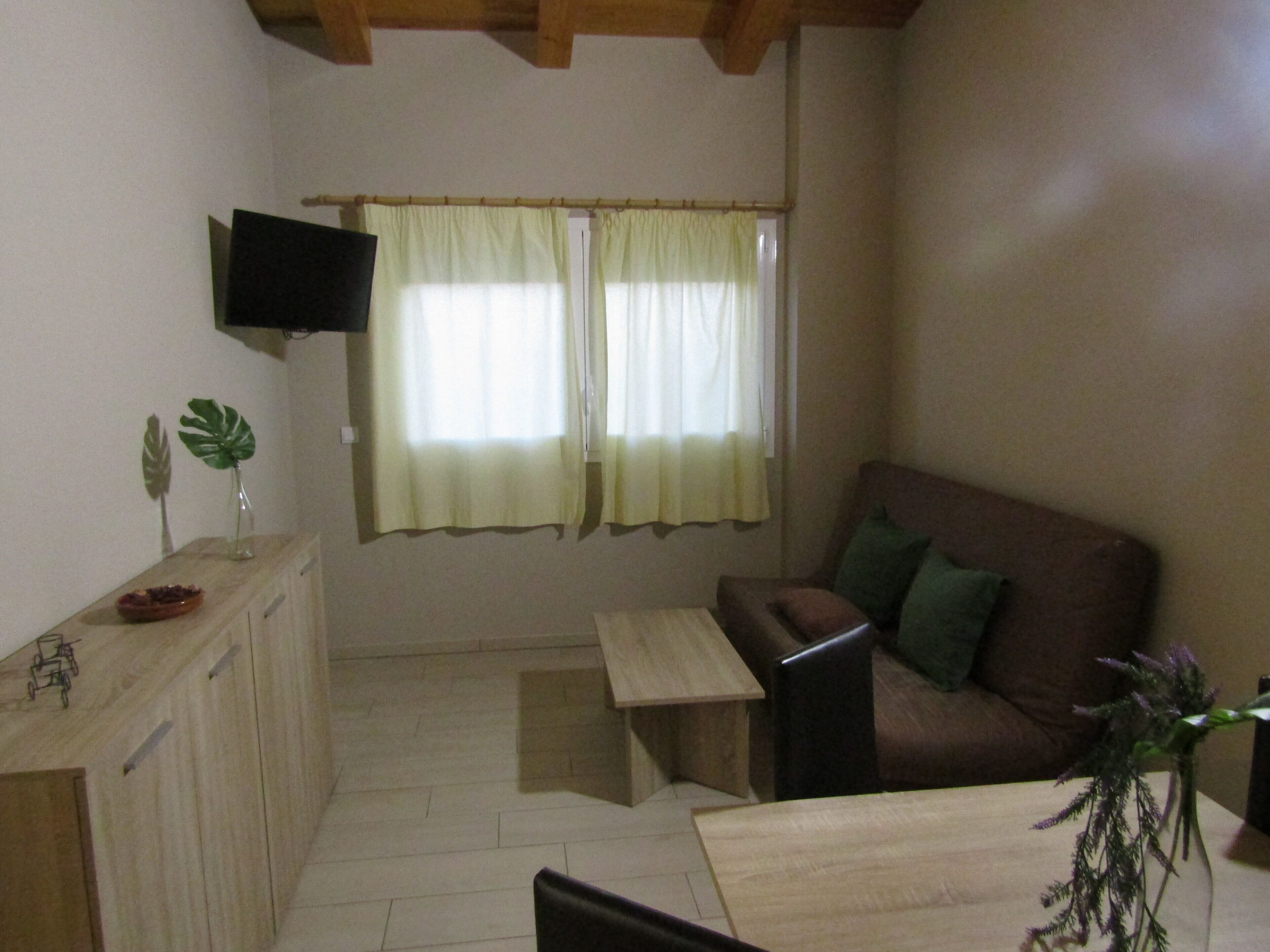 A small, cozy living room with a wooden floor, a brown sofa, a small table, a TV mounted on the wall, and two windows with light curtains.