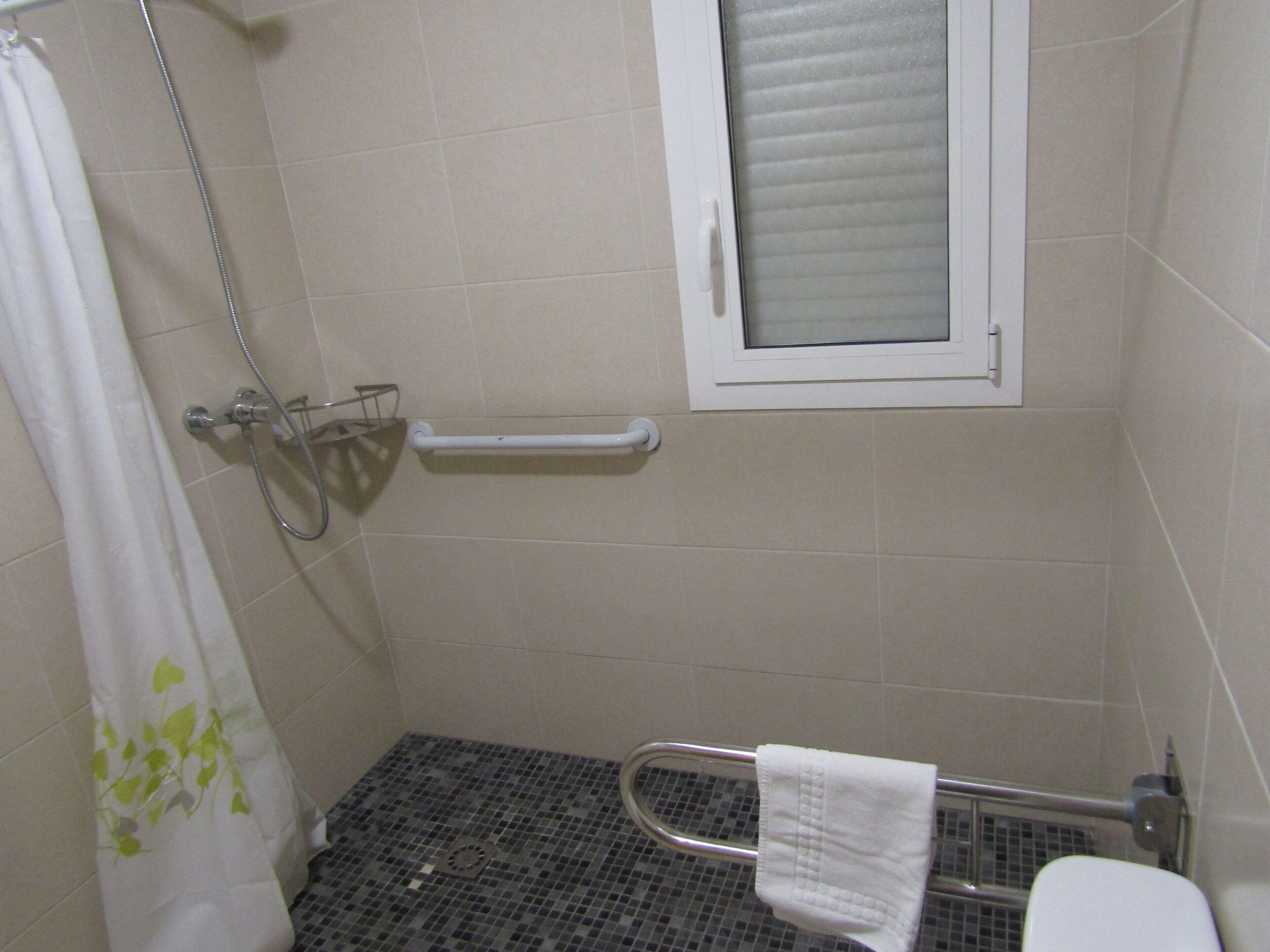 A small, tiled bathroom with a shower, a handrail, a window, and a towel rack.