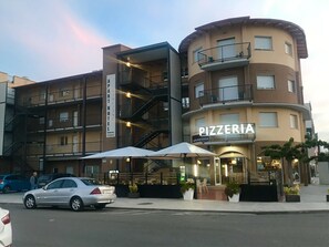 A multi-story building with a 'PIZZERIA' sign, a 'GUILIANO PART HOTEL' sign, and a parking area with cars. - Aparthotel Giuliano (Castello d'Empuries)