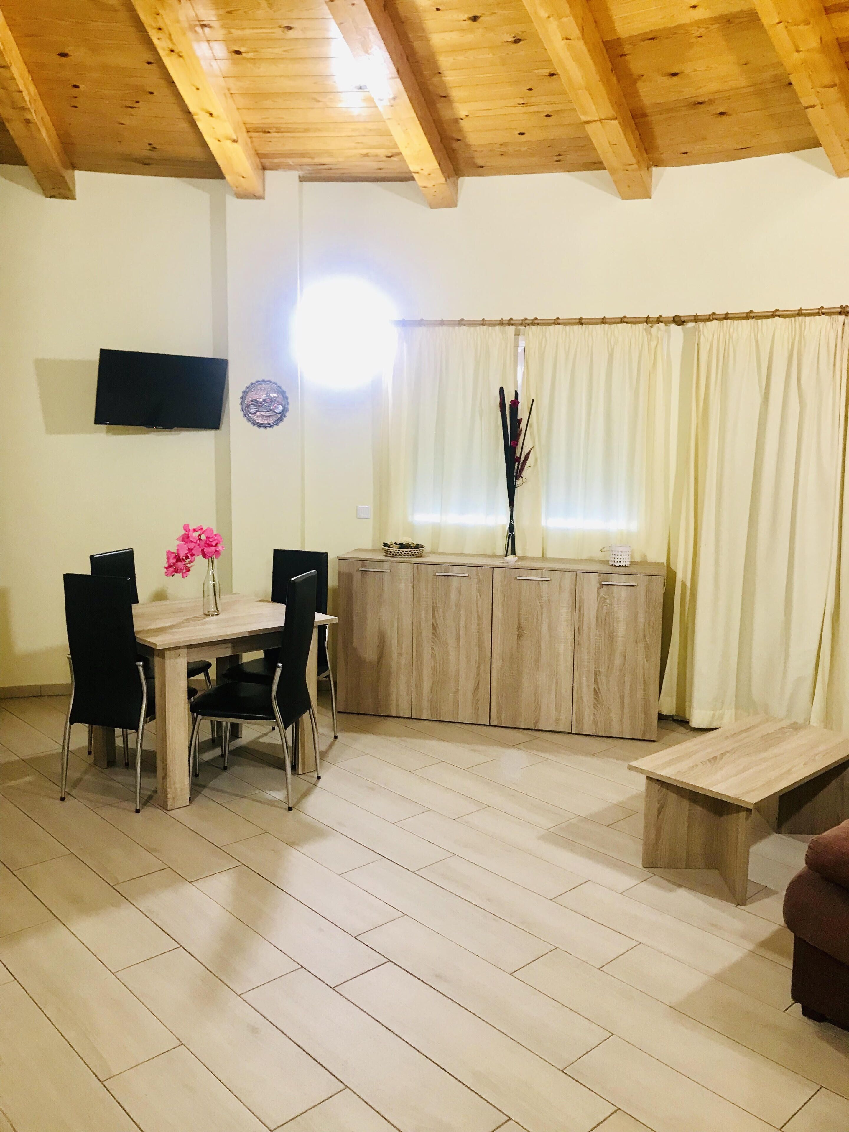 Een eetgedeelte met een houten tafel en stoelen, een bank, een aan de muur bevestigde televisie en een raam met gordijnen.