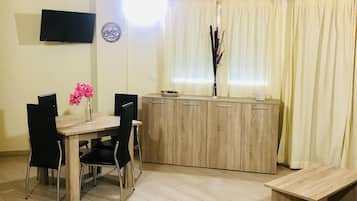 Uma área de refeições com mesa e cadeiras de madeira, um banco, uma televisão montada na parede e uma janela com cortinas.
