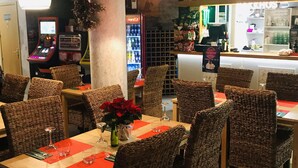 Interior de restaurante con sillas de mimbre, mesas con manteles individuales rojos, una zona de bar con botellas y un refrigerador Coca-Cola.