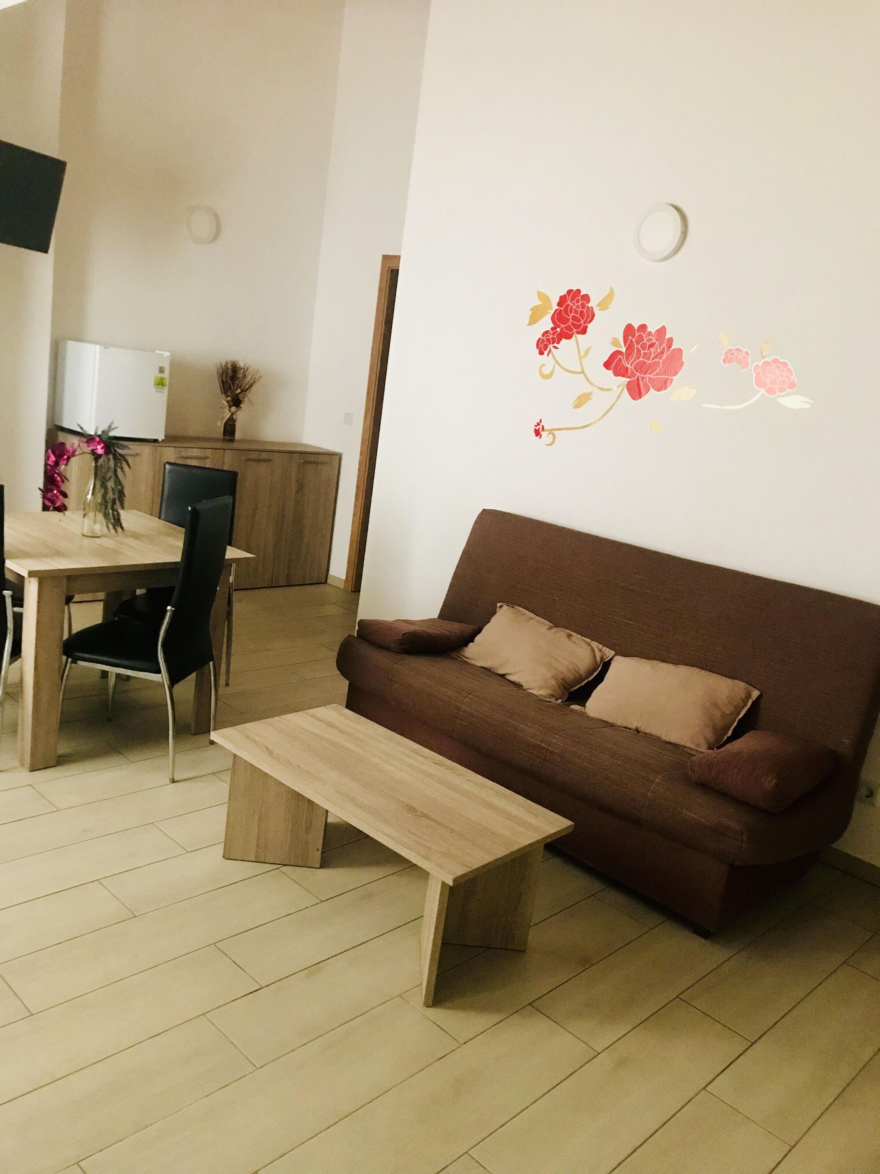 A living room with a brown sofa, a wooden coffee table, and a dining area with a white refrigerator and chairs.