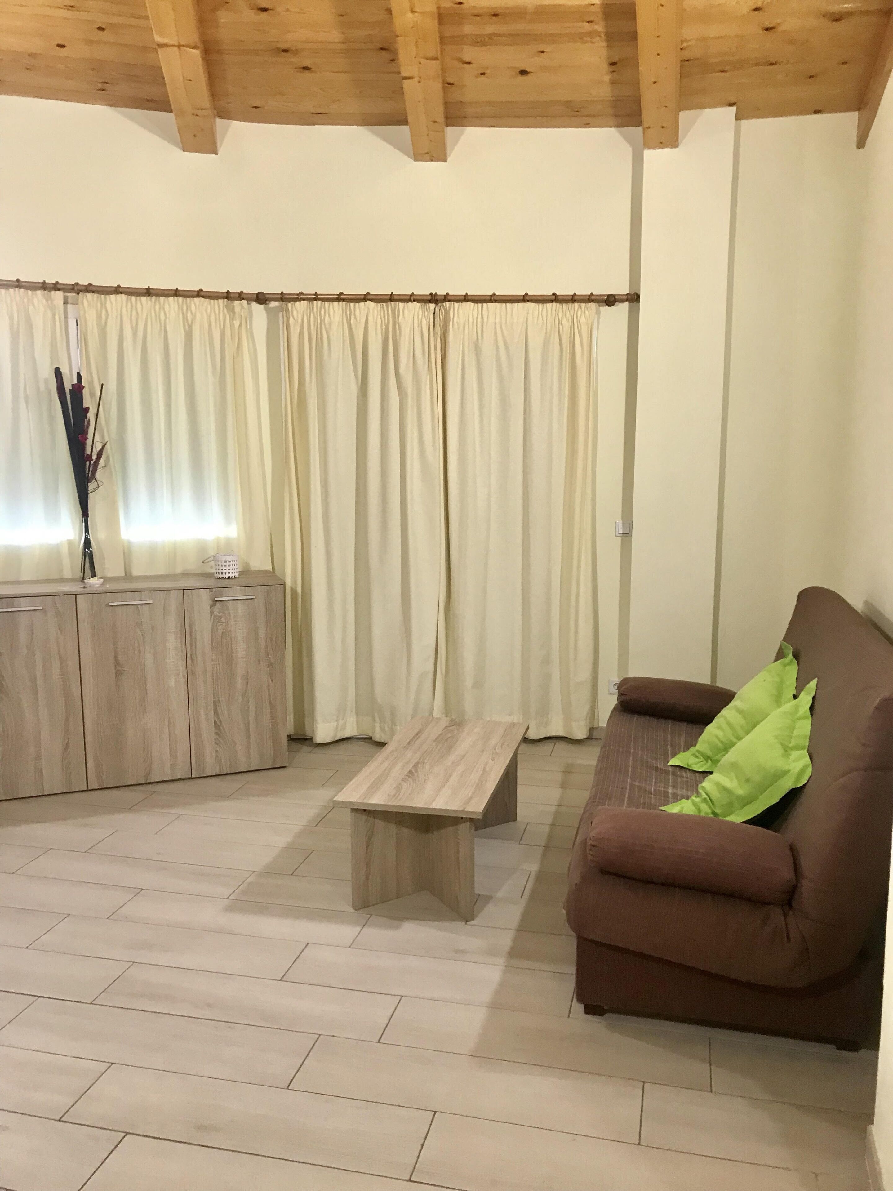 Een woonkamer met een houten plafond, een bruine bank met een groen kussen, een houten salontafel en een kastje met een vaas en kaars.
