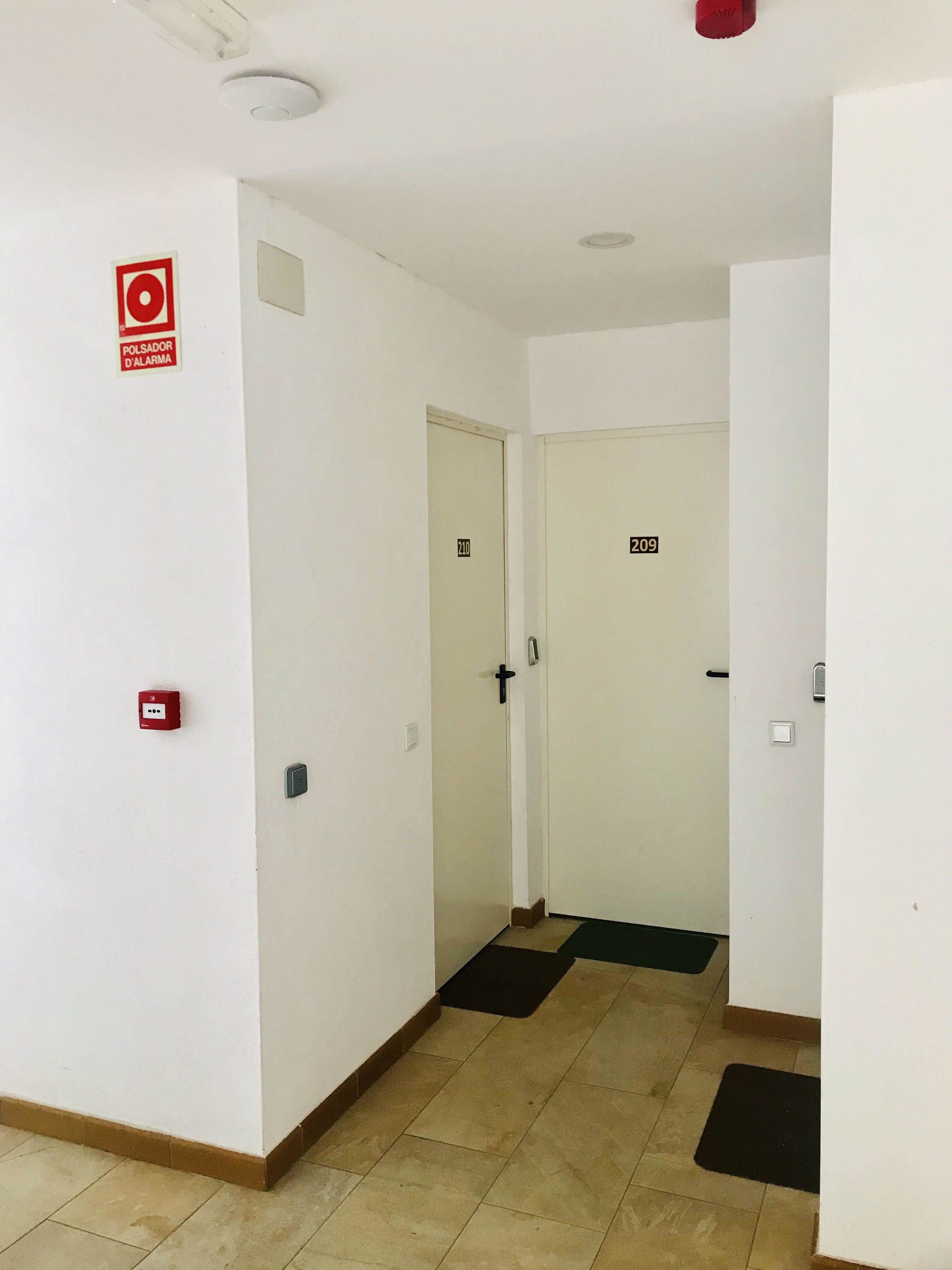 Un corridoio con pareti bianche, porte numerate, un pulsante rosso di allarme antincendio e un cartello di allarme.