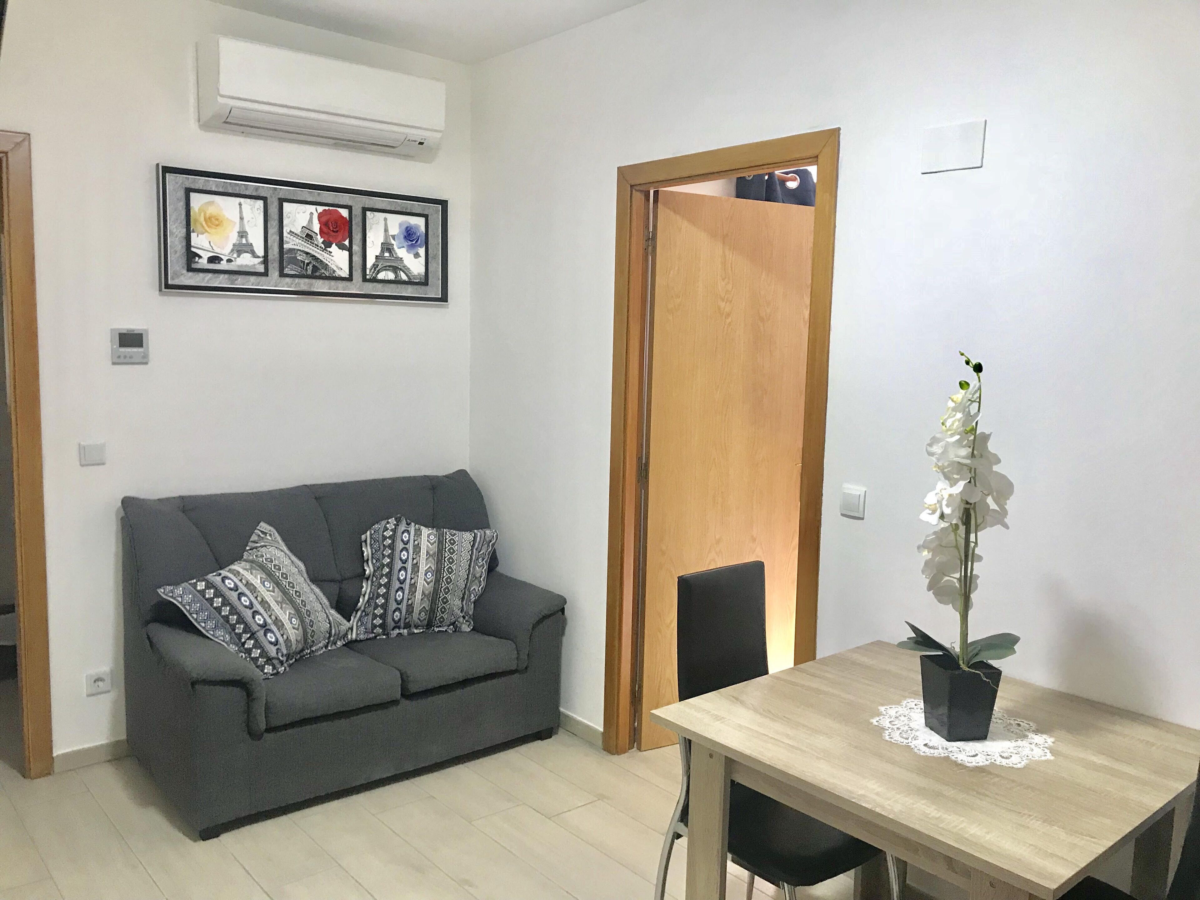Una sala de estar con un sofá gris, cojines estampados, una mesa de comedor de madera con una orquídea blanca y una puerta con un espejo.